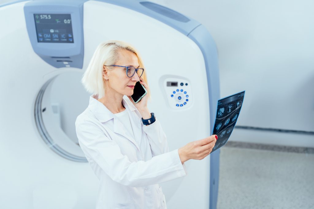 Portrait of cheerful blond woman doctor at modern tomography room having phone conversation and looking X-ray image with CT scanner on background.