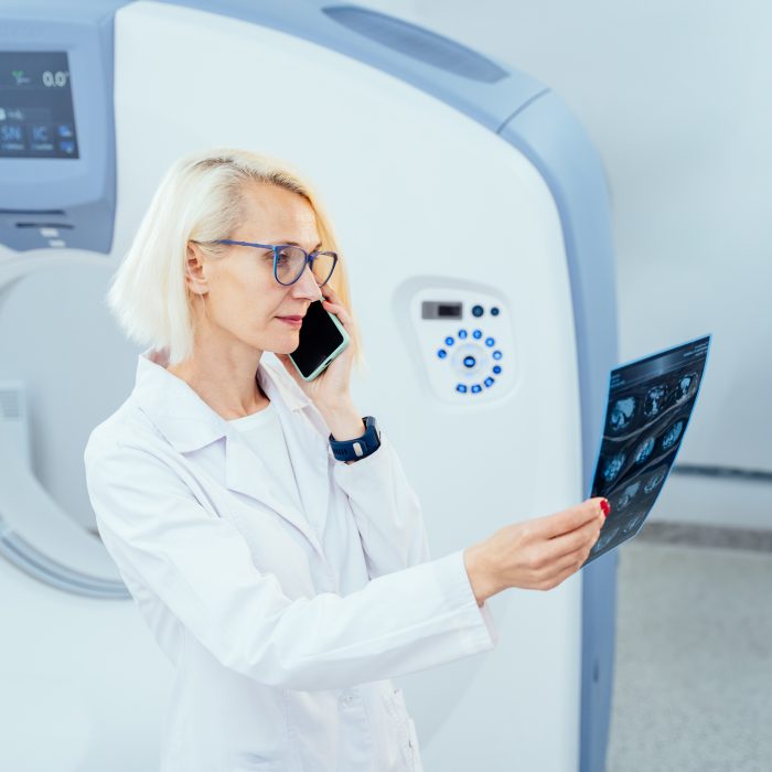 Portrait of cheerful blond woman doctor at modern tomography room having phone conversation and looking X-ray image with CT scanner on background.