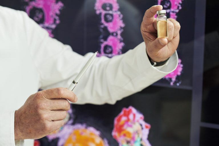 Hand of middle-aged male scientist holding vial with yellow liquid and pen, standing in front of medical imaging scans, demonstrating scientific or medical research at conference.