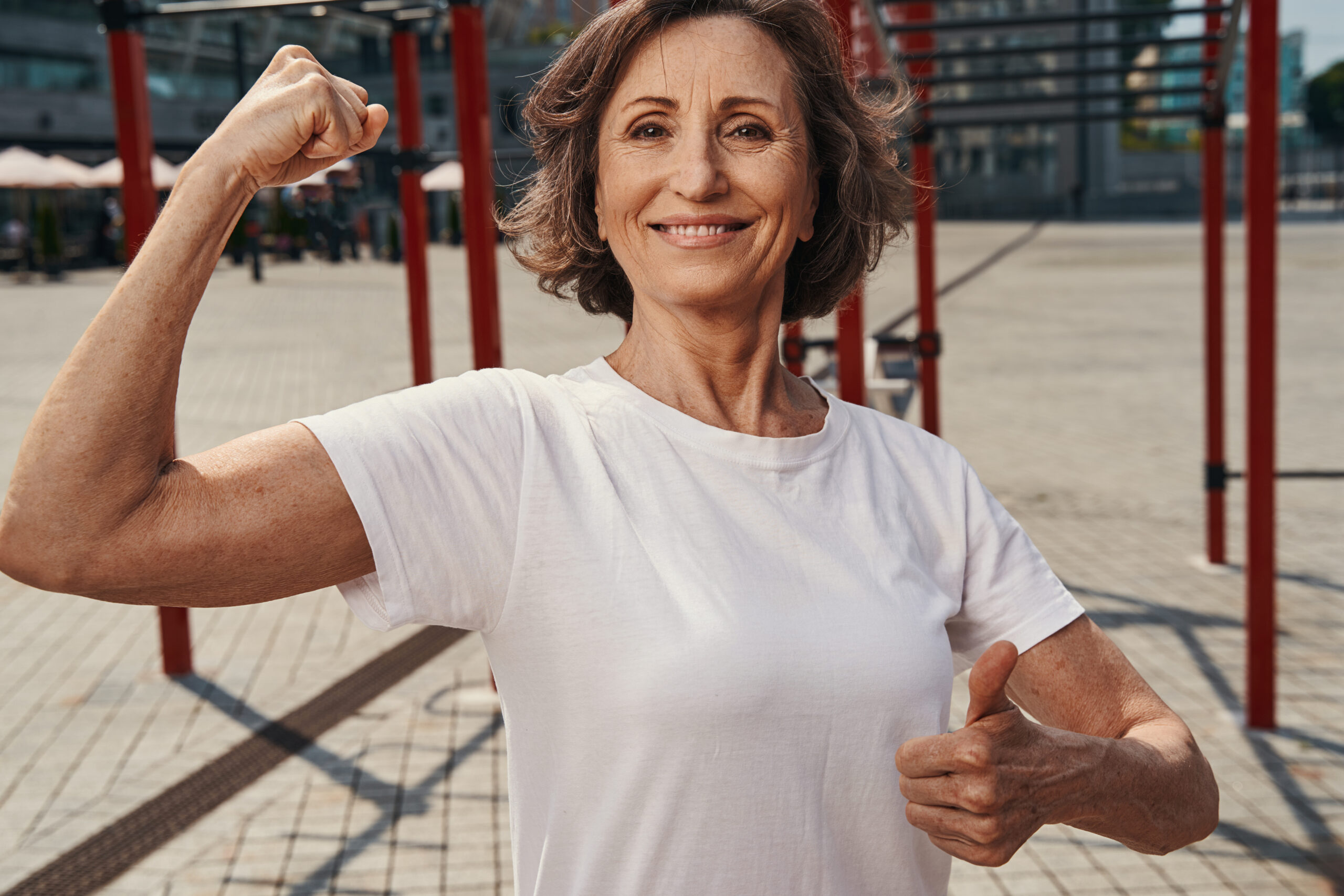 Waist up portrait of smiling mature female showing trained biceps and thumb up near horizontal bars.