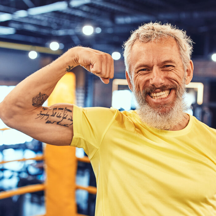 Strong and fit at any age. Portrait of a happy handsome mature man showing his biceps and smiling at camera while standing on the boxing ring at gym. Sport, box, fitness, healthy and active lifestyle.